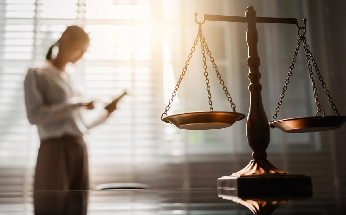 The scales of justice on a desk with a lawyer reading documents by a bright window behind